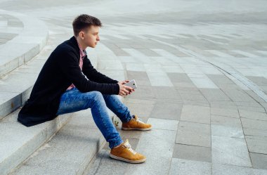 Ekose gömlek ve kot pantolon ile smartphone merdivenlerde el açık havada oturup şık genç adam. Öğrenci ile mesafe içine boş alan seyir smartphone