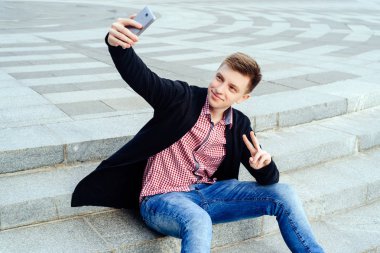 Şık genç gülümseyen adam ekose gömlek ve merdivenlerde oturan ve selfie yapım ve iki başparmak gösteren kot açık havada jest. Teknoloji ve iletişim kavramı 