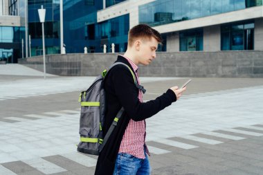 Şehirde yürüyüş ve açık havada smartphone kullanarak sırt çantası ile ekose gömlekli şık genç adam. Öğrenci ile yürüyüş smartphone 