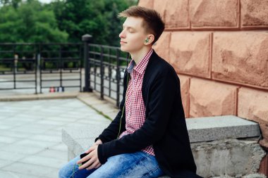 Sessiz sakin genç adam ekose gömlek ve ceket cep telefonu açık havada kulaklık ile müzik dinlemek. Akıllı telefon ve kulaklık ile oturan kapalı gözler ile öğrenci