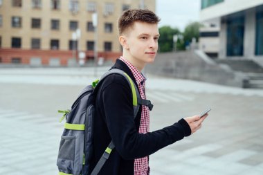 Şehirde yürüyüş ve açık havada smartphone kullanarak sırt çantası ile ekose gömlekli şık genç adam. Cep telefonu üzerinde street yürüyüş ile öğrenci