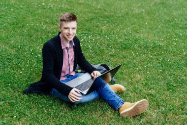 Yong okuma, dizüstü bilgisayarda çalışan adam ekose gömlek ve ceket kampüste, yeşil çim üzerinde oturan gülümsüyor. Dizüstü bilgisayar açık havada diz ile öğrenci