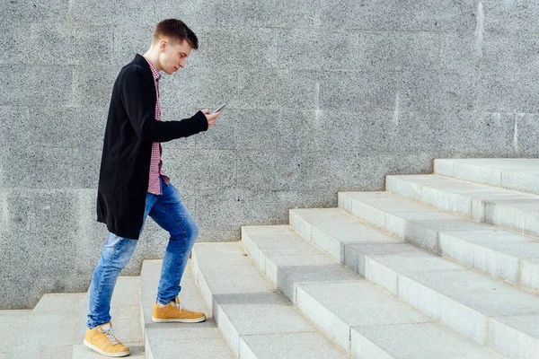 Ekose gömlek ve merdivenlerden yukarı yürümek ve açık havada smartphone kullanarak kot şık genç adam. Üst katta, boş alan yürüyüş smartphone ile öğrenci