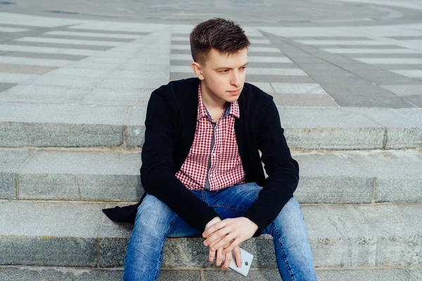 Ekose gömlek ve kot pantolon ile smartphone merdivenlerde el açık havada oturup şık genç adam. Öğrenci ile mesafe içine boş alan seyir smartphone