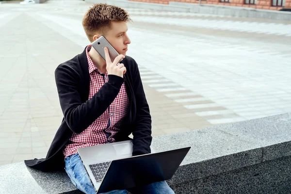 Şehirde oturan ve dizüstü bilgisayar açık havada diz ile cep telefonuyla konuşurken ekose gömlekli şık genç adam. Cep telefonu ve laptop ile öğrenci