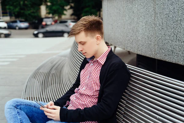 Şehirde bankta oturmuş akıllı telefon kullanan rahat genç adam. Holding ve seyir mobil akıllı telefon açık havada ceketli yakışıklı öğrenci