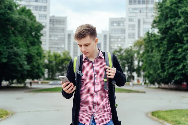 Şehirde yürüyüş ve açık havada smartphone kullanarak sırt çantası ile ekose gömlekli rahat genç adam. Cep telefonu üzerinde street yürüyüş ile öğrenci