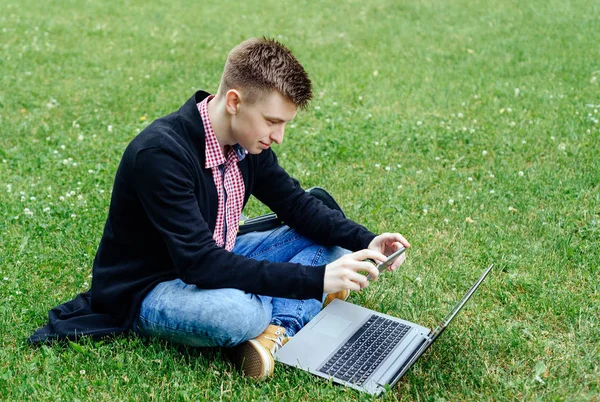Genç gülümseyen adam ekose gömlek ve yeşil çimenlerin üzerine oturmuş ve dizüstü bilgisayarda çalışırken cep telefonu kullanarak ceket. Öğrenci dizüstü bilgisayar ile açık havada