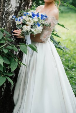 Güzel gelin düğün elbise düğün buket peonies, mavi çiçek ve yeşillik ayakta açık havada huş yakınındaki