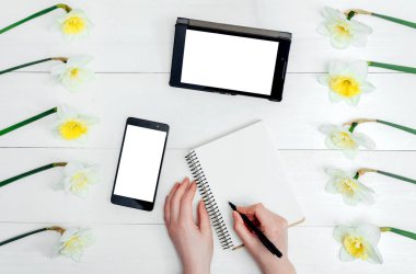 Üstten görünüm nergis, boş günlüğü, tablet, telefon ve kızın elleri kalem, beyaz ekran, boş alan ile tablo. Kızın ellerini günlüğüne yazma