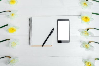 Beyaz ahşap zemin üzerine sarı nergis çerçevesinde. Boş kart düz yatıyordu. Nergis, boş günlüğü, kalem ve telefon, boş alan, beyaz ekran ile tabloda üstten görünüm