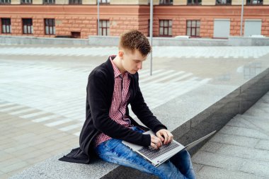 Oturma ve açık havada, diz dizüstü bilgisayarda yazarak kot pantolon ve ceket şık genç adam boş alan. Teknoloji ve iletişim kavramı 