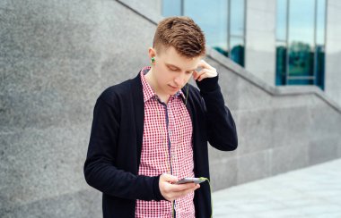 Neşeli mutlu genç adam ekose gömlek ve ceket cep telefonu açık havada kulaklık ile müzik dinlemek. Akıllı telefon ve kulaklık, boş alan ile öğrenci