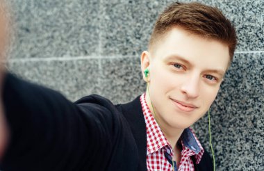 Selfie fotoğraf çekici gülümseyen genç adam ekose gömlek ve ceket ile kulaklıklar. Akıllı telefon müzik öğrenci