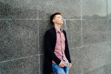 Neşeli mutlu genç adam ekose gömlek ve ceket cep telefonu açık havada kulaklık ile müzik dinlemek. Akıllı telefon ve kulaklık, boş alan ile öğrenci