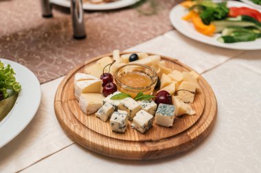 Peynir tabağı kiraz ve bal ile yuvarlak ahşap tahta plaka kutlama yemek masası, seçici odak üzerinde. Camembert, Dorblu, Emmental, Brie kesme tahtası üzerinde