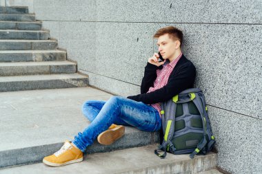 Yakışıklı delikanlı taş merdiven ve şehir havada cep telefonuyla konuşurken yakın oturan sırt çantası ile boş alan. Öğrenci üniversite açık havada sonra aittir