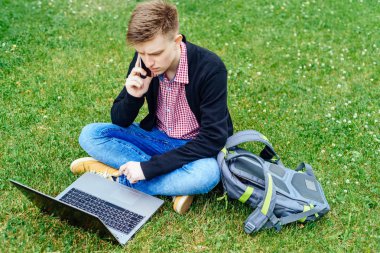 Melankoli delikanlı yeşil çim çim üzerinde oturan ve yaz aylarında açık havada dizüstü bilgisayarda çalışırken cep telefonuyla konuşan sırt çantası ile. Öğrenci dizüstü bilgisayar ile boş alan