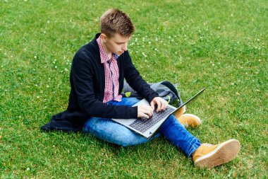 Okuma, dizüstü bilgisayarda çalışarak Yong adam ekose gömlek, ceket ve kot kampüste, yeşil çim üzerinde oturuyor. Öğrenci dizüstü bilgisayar açık havada, diz ile boş alan