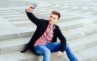 Şık genç gülümseyen adam ekose gömlek ve merdivenlerde oturan ve açık havada, selfie yapma kot boş alan. Teknoloji ve iletişim kavramı 