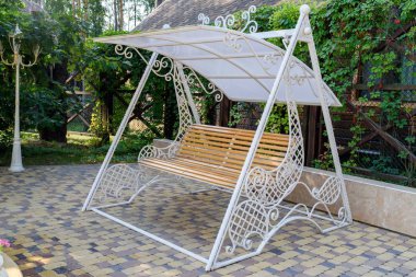 Empty wooden swing with white decorative metal frame in the garden, free space. Wooden furniture for garden. Swinging bench in the garden clipart