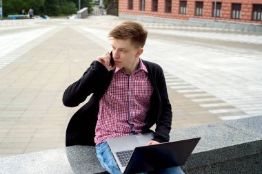Taş açık dizüstü bilgisayar ile oturup ve akıllı telefon şehir havada konuşurken yakışıklı bir genç adam. Öğrenci üniversiteden sonra açık havada, boş alan aittir.