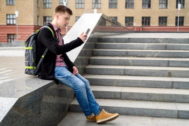 Yakışıklı delikanlı taş merdiven yanında oturan ve akıllı telefon şehir açık, tam vücut kullanarak sırt çantası ile. Öğrenci üniversite açık havada sonra aittir