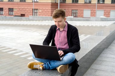 Oturma ve açık havada, diz dizüstü bilgisayarda yazarak kot pantolon ve ceket şık genç adam boş alan. Teknoloji ve iletişim kavramı 