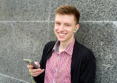 Neşeli mutlu genç adam ekose gömlek ve ceket cep telefonu açık havada kulaklık ile müzik dinlemek. Akıllı telefon ve kulaklık, boş alan ile öğrenci