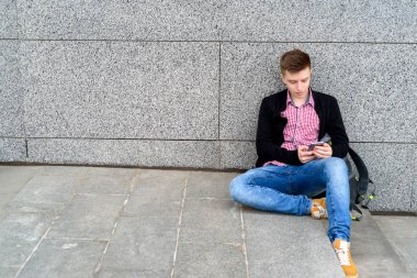 Yakışıklı delikanlı taş duvara yakın oturan ve şehir mekanlarda cep telefonu kullanma sırt çantası ile boş alan. Öğrenci üniversite açık havada sonra aittir
