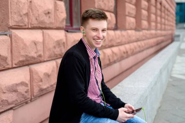 Neşeli mutlu genç adam ekose gömlek ve ceket cep telefonundan kulaklık ile müzik dinlemek ve açık havada şarkı. Akıllı telefon ve kulaklık ile öğrenci