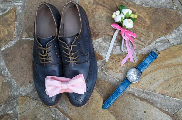 Top view of blue leather groom shoes, watches, boutonniere and pink bowtie on brown natural stone. Groom wedding accessories. Man watches, bow-tie, boutonniere and footwear on stone background