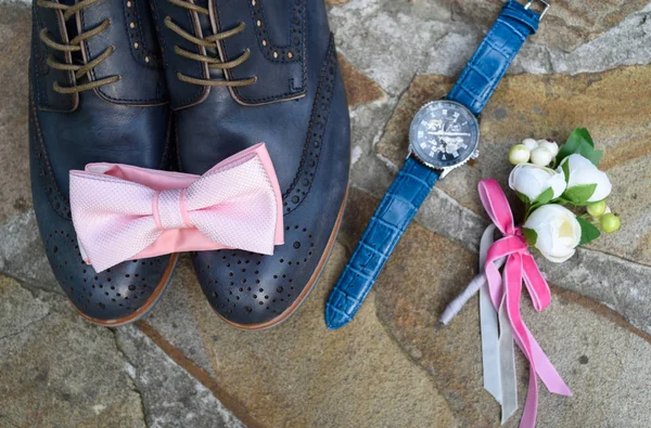 Top view of pink bowtie on blue leather groom shoes near watches and boutonniere on brown natural stone. Groom wedding accessories. Man watches, bow-tie, boutonniere and footwear on stone background