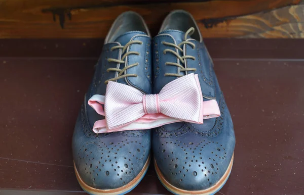 Close up of pink bowtie on blue leather shoes. Modern man accessories. Wedding details. Groom accessories. Men's casual outfits with shoes and bowtie on wooden background