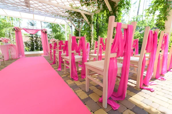 Empty wedding chairs on each side of archway. Place for wedding ceremony decorated in pink color,  wooden chairs for guests outdoors. Wedding ceremony in pink color