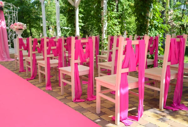 Wedding chairs on each side of archway. Place for wedding ceremony decorated in pink color,  wooden chairs for guests outdoors. Wedding ceremony in pink color