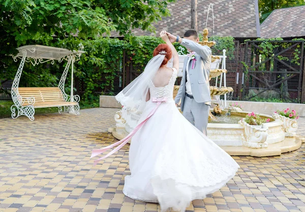 Happy groom and beautiful redhead bride in white dress holding hands and dancing outdoors, free space. Beautiful wedding couple spinning in wedding day. Wedding dress with bow on back flutters in the wind
