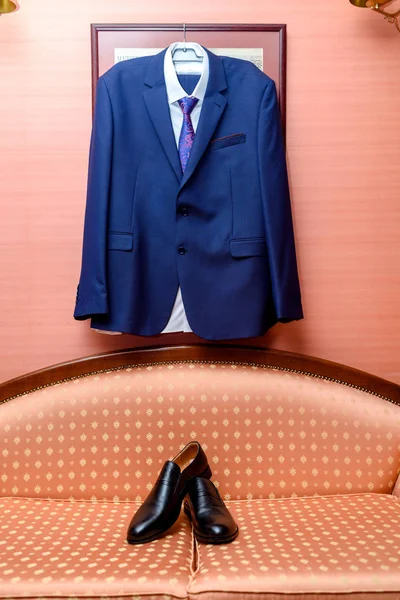 Dark blue suit, white shirt and necktie hanging on the wall over black leather shoes. Groom clothing indoors. Set of classic man clothes. Groom accessories. Shoes on the sofa under groom hanging suit