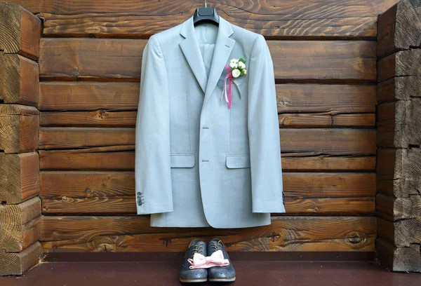 Stylish elegant wedding groom suit with buttonhole hanging on wooden background. Gray suit hangs above leather groom shoes and pink bowtie. Groom wedding accessories, free space
