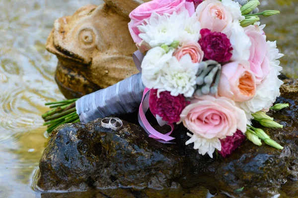Close up of two golden wedding rings on stone in water near wedding bouquet in fountain, free space. Wedding details outdoor with copy space