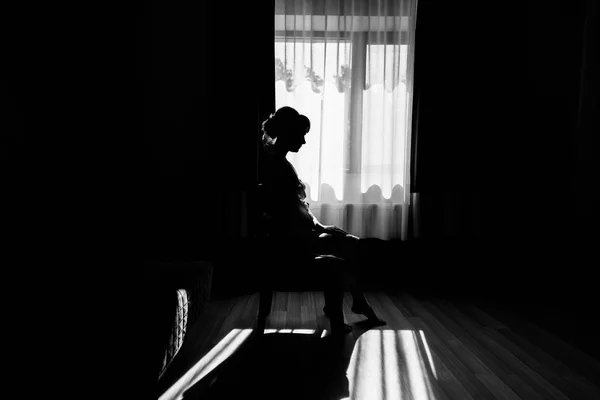 Silhouette of beautiful bride sitting on chair at the window in bedroom. Morning of the bride. Bride in gown posing in sunlight at window in morning. Getting ready. Black and white photo