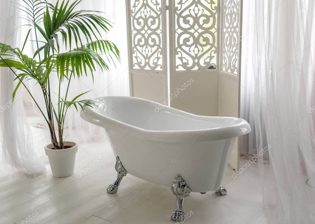 Beautiful luxury vintage empty bathtub near big window in bathroom interio, free space. Freestanding white bath near folding screen and palm tree, copy space 