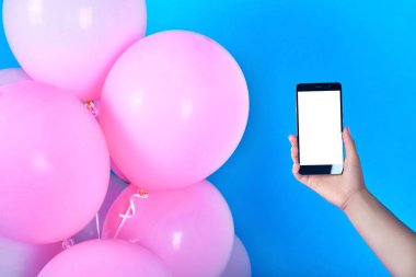 Mavi arka plan üzerinde pembe balonlar. El, kopya alanı boş beyaz ekran ile mobil akıllı telefon. Pembe pastel parti balonu, boş alan. Aşk, mutluluk, Sevgililer günü, düğün, tatil kavramı