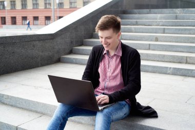 Merdivenlerde oturan ve açık havada dizüstü bilgisayarda yazarak kot pantolon ve ceket genç adam gülümseyerek mutlu. Teknoloji ve iletişim kavramı 