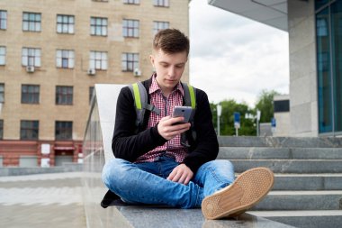 Yakışıklı delikanlı taş merdiven yanında oturan ve akıllı telefon şehir açık, tam vücut kullanarak sırt çantası ile. Öğrenci üniversite açık havada sonra aittir