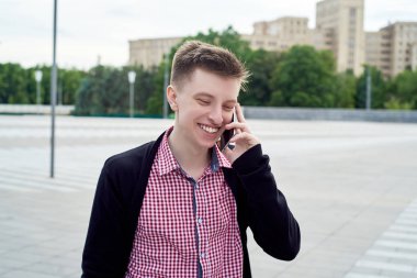 Ekose gömlek ve ceket şehirde yürüyüş ve cep telefonu açık havada üzerinde konuşmak mutlu gülümseyen genç adam boş alan. Mobil akıllı telefon ile öğrenci