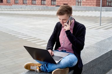 Taş açık dizüstü bilgisayar ile oturup ve akıllı telefon şehir havada konuşurken mutlu gülümseyen genç adam. Öğrenci üniversiteden sonra açık havada, boş alan aittir.