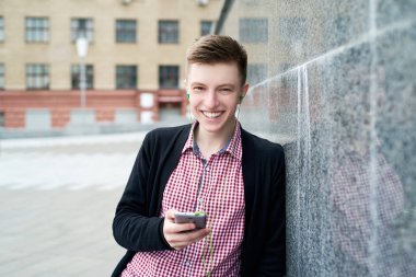 Neşeli mutlu genç adam ekose gömlek ve ceket gri taş duvar arka plan açık havada mobil akıllı telefon kulaklık ile müzik dinleme boş alan