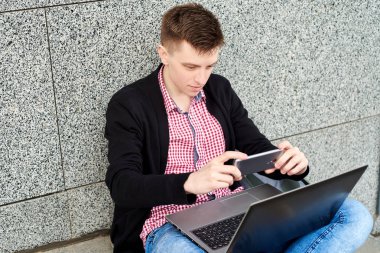 Dizüstü bilgisayar ile taş duvara yakın oturan ve tarama Qr kodu mobil akıllı telefon ile genç adam portresi. Öğrenci cep telefonu uygulaması barkod tarama kullanarak. Online alışveriş, teknoloji 