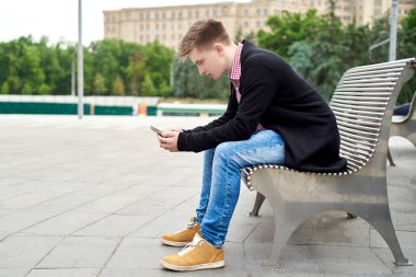 Şehir sokak, kopya alanı banktaoturuyor smartphone ile genç adam. Cep telefonu kullaranarak ve ekran açık, yan manzaraya bakarak Genç adam portresi. İnsanlar, teknoloji ve seyahat kavramı